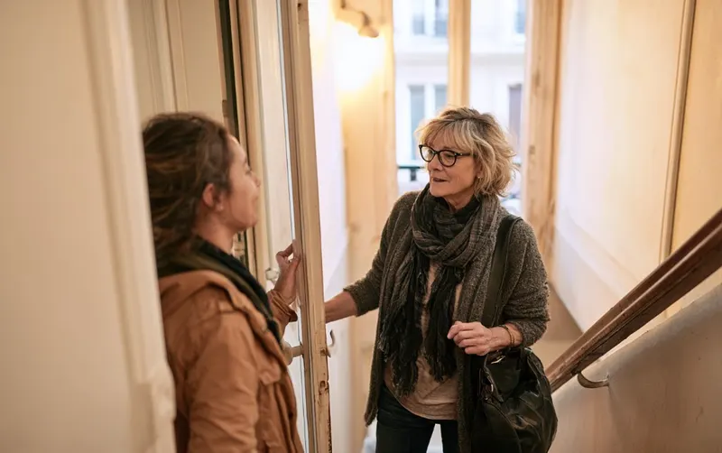 Deux voisines parlent sur le palier d'un immeuble français, porte entrebâillée, lumière de fin d'après-midi