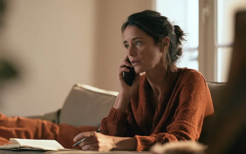 Une personne appelle depuis son canapé, combiné à l'oreille, carnet ouvert sur la table